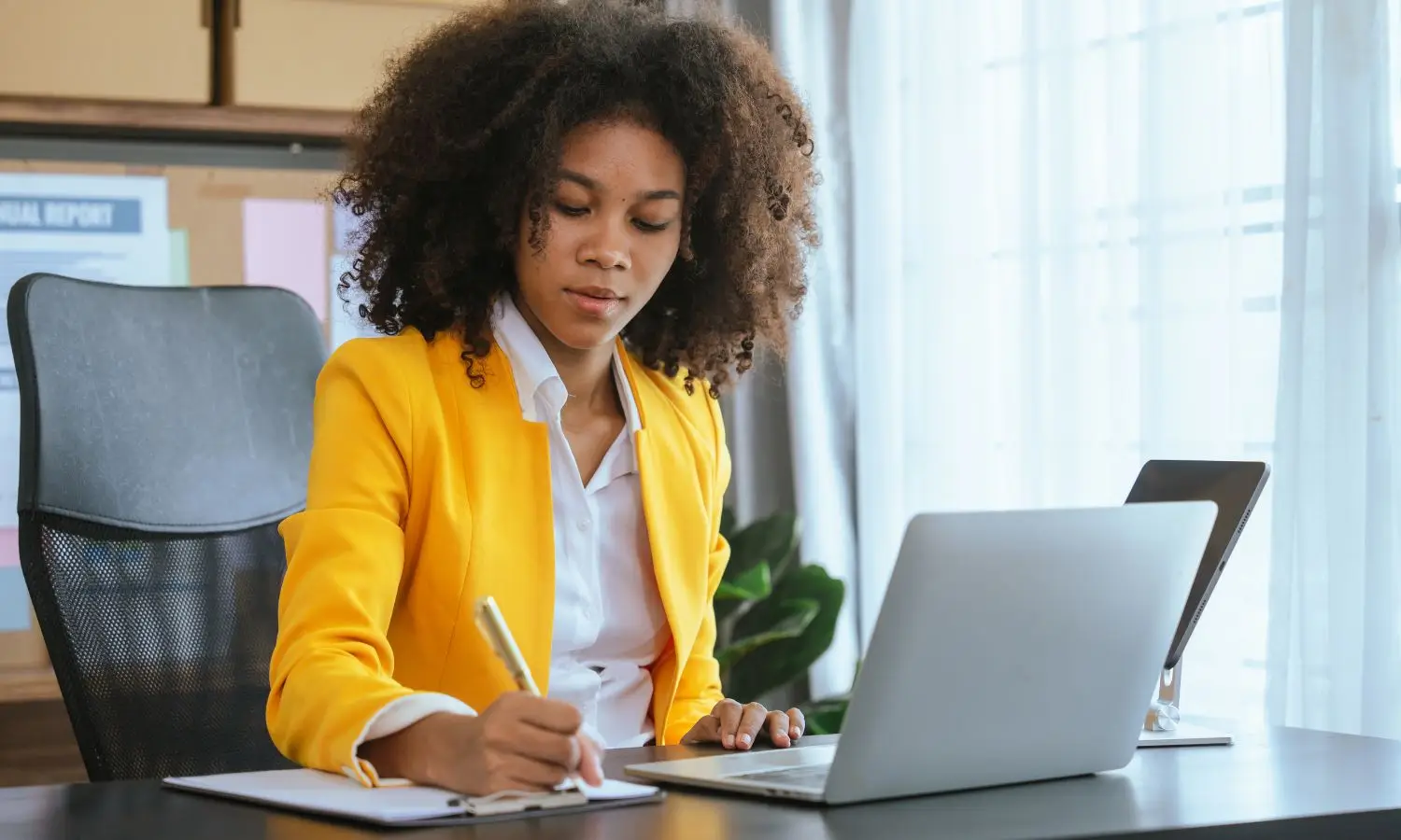 African American woman with natural curly loose afro wearing a yellow jacket and white blouse. She's siting at a desk in front of a laptop taking notes while watching laptop.